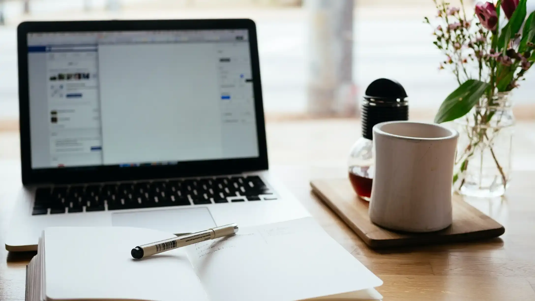 A laptop displaying a Facebook page sitting on a table next to a notepad and mug