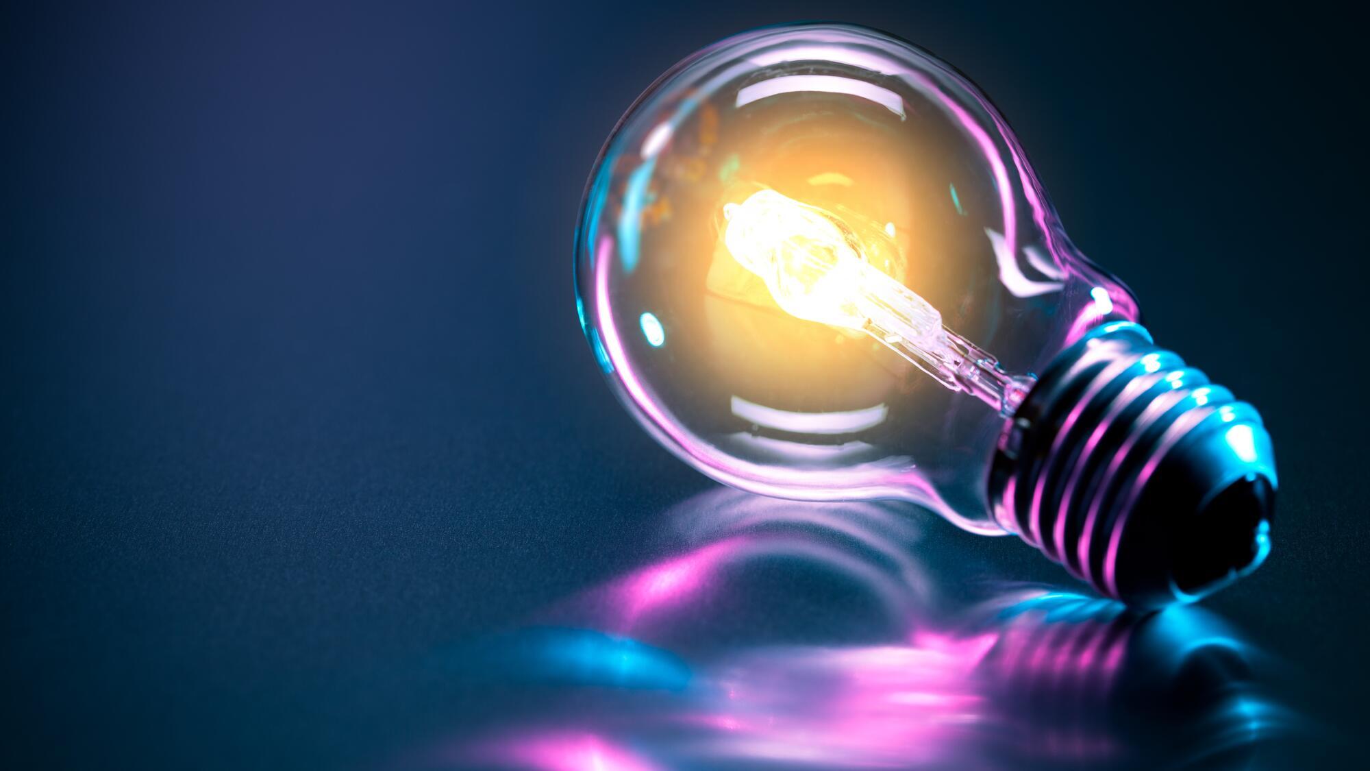 A lit up lightbulb lays on a reflective surface, casting a purple-tinted reflection