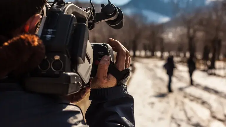 In the foreground a man holds a camera, filming two people in the distance near some trees, there is a mountain in the background