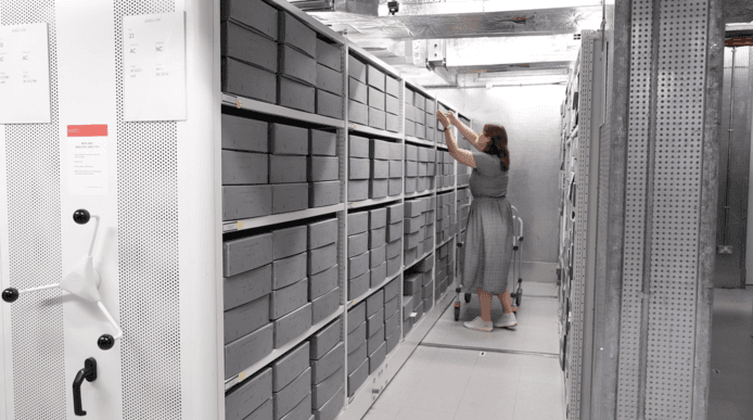 A woman in a room filled with archived folders, taking one of them out.