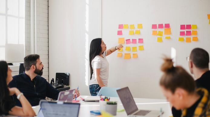 A woman stands in an office in front of a wall of post-it notes, people are sat around a meeting table in the foreground