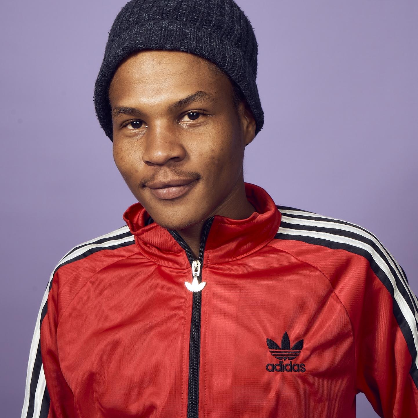 Innocent, a young man in a beanie hat and red Adidas tracksuit top standing in front of a lilac wall  