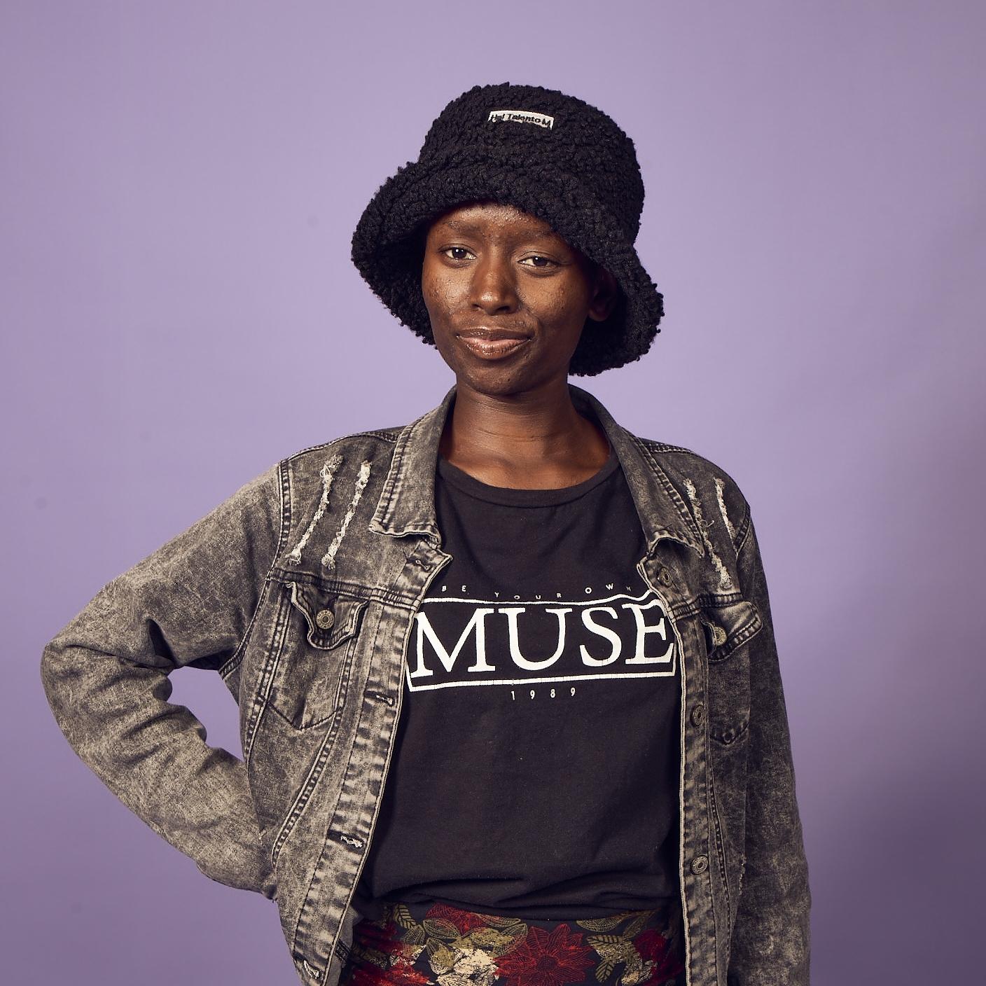 Winnie, a young women in a black bucket hat, grey jacket, black t-shirt and patterned leggings stands in front of a lilac wall