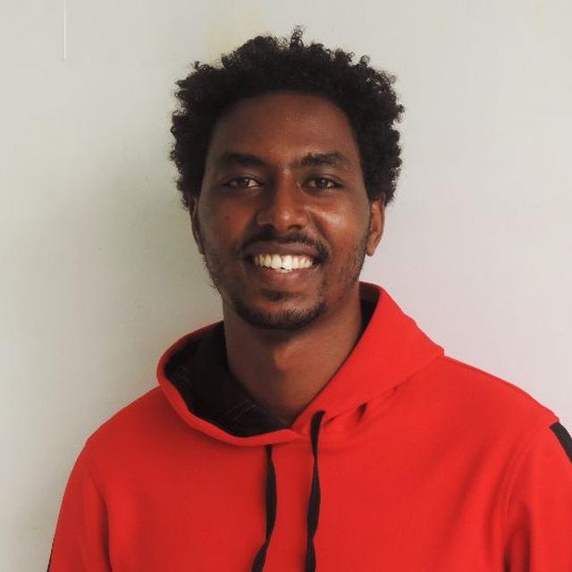 Habteab, a young man stands in front of a white wall in a red hoodie, smiling