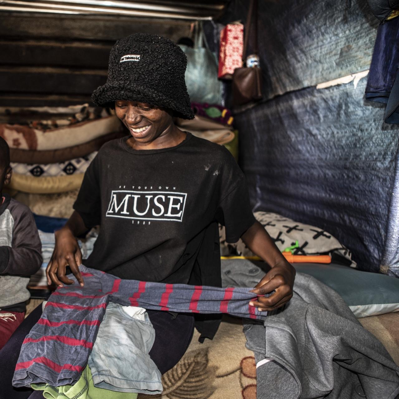 Winnie, a young women in a black bucket hat and black t-shirt sits next to young boy (her son) on a bed, folding clothes