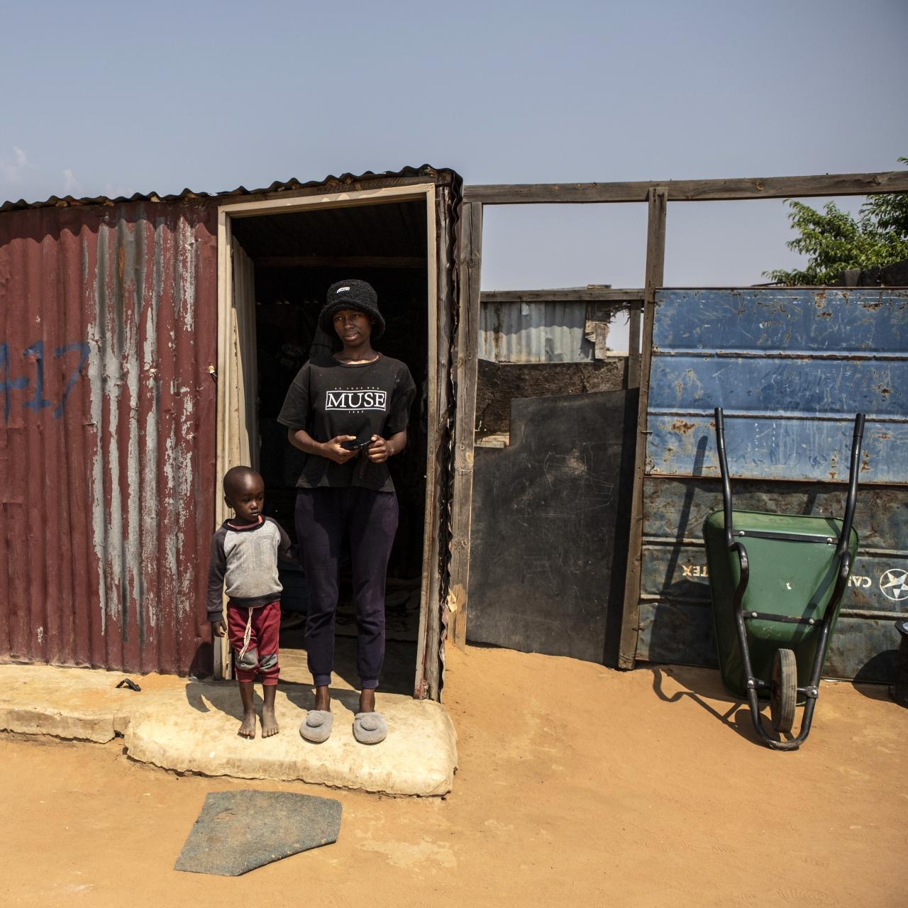 Winnie, a young women in a black bucket hat and black t-shirt stands next to young boy (her son) in the doorway of a small corrugated iron house