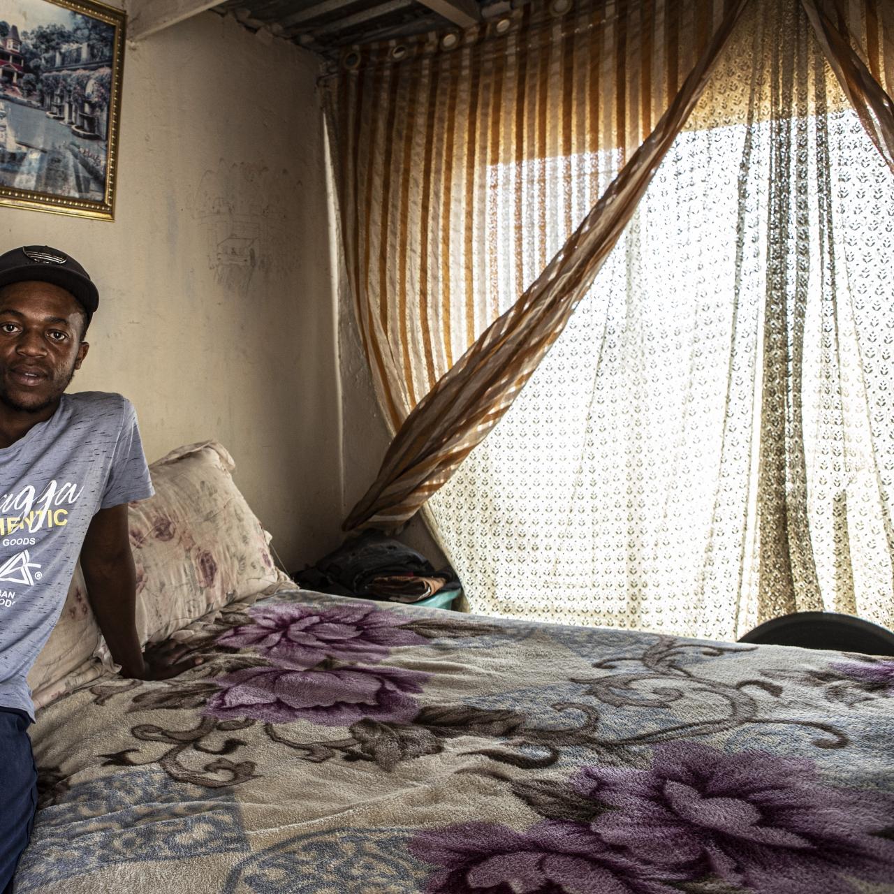 Bheki, a young man in a cap and light blue t-shirt sits on a double bed with floral sheets