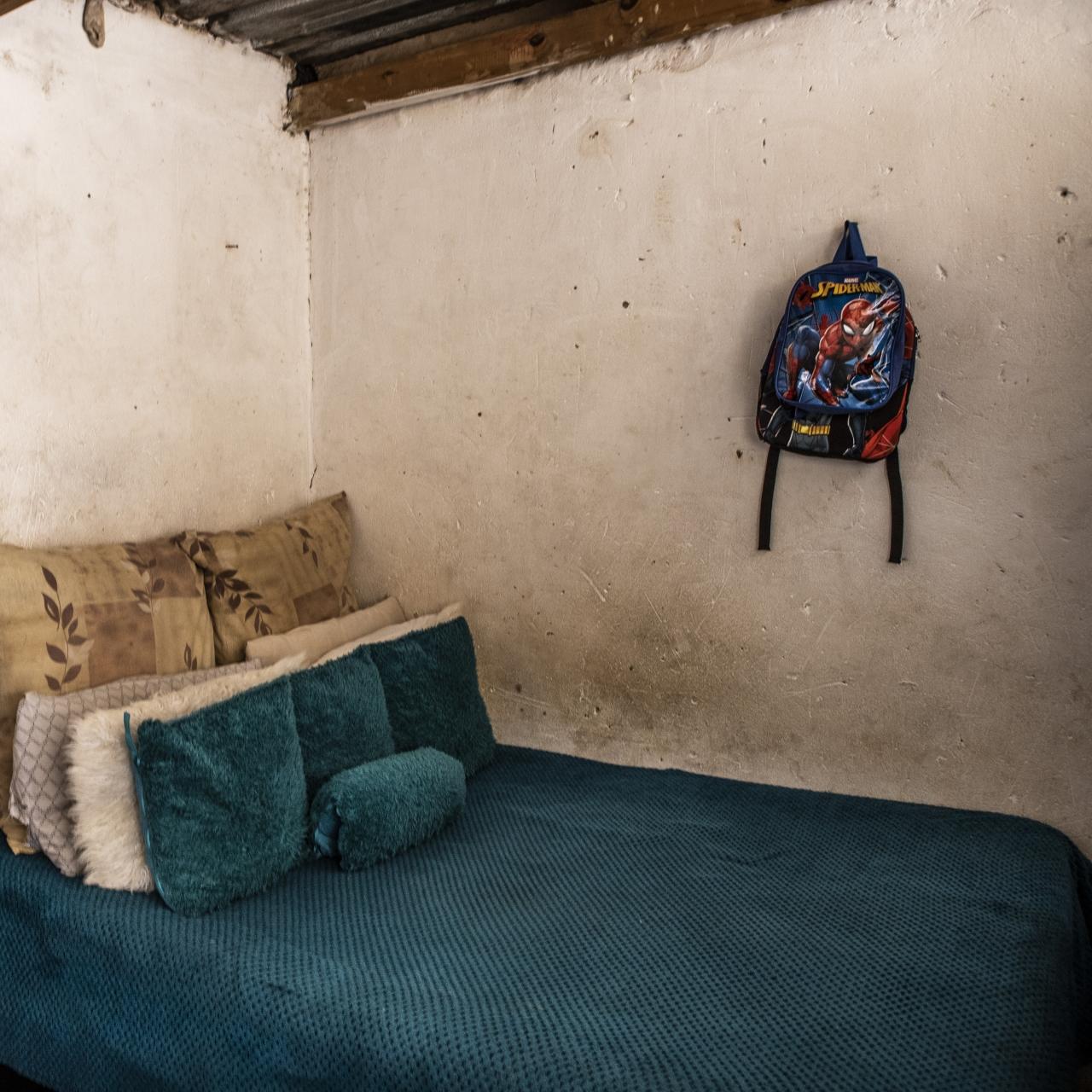 A small bedroom with rough, white walls and a bed with turquoise sheets. A Spiderman backpack hangs on the wall and large stereo sits atop a bedside table