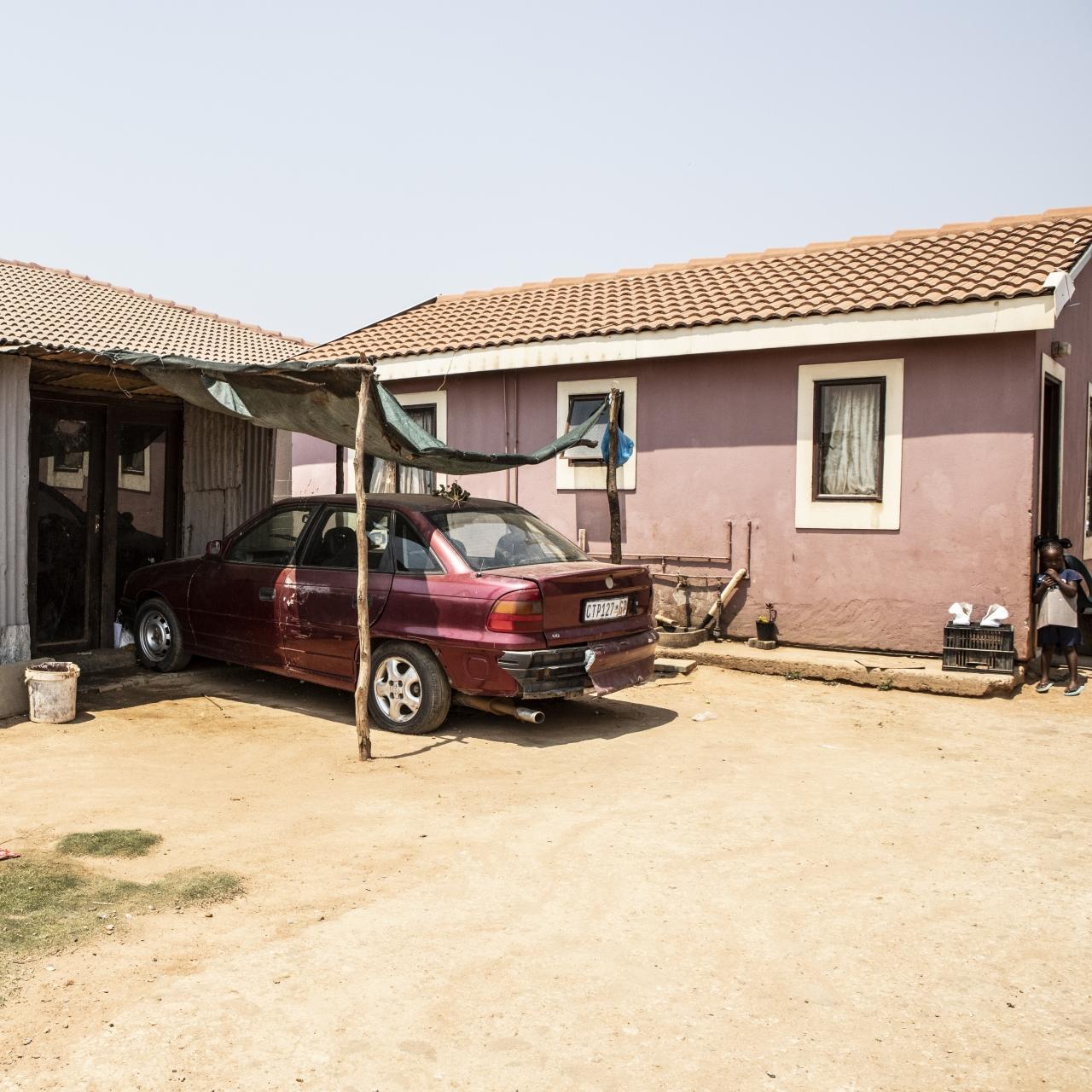 A red car parked under a makeshift car port outside two adjacent houses
