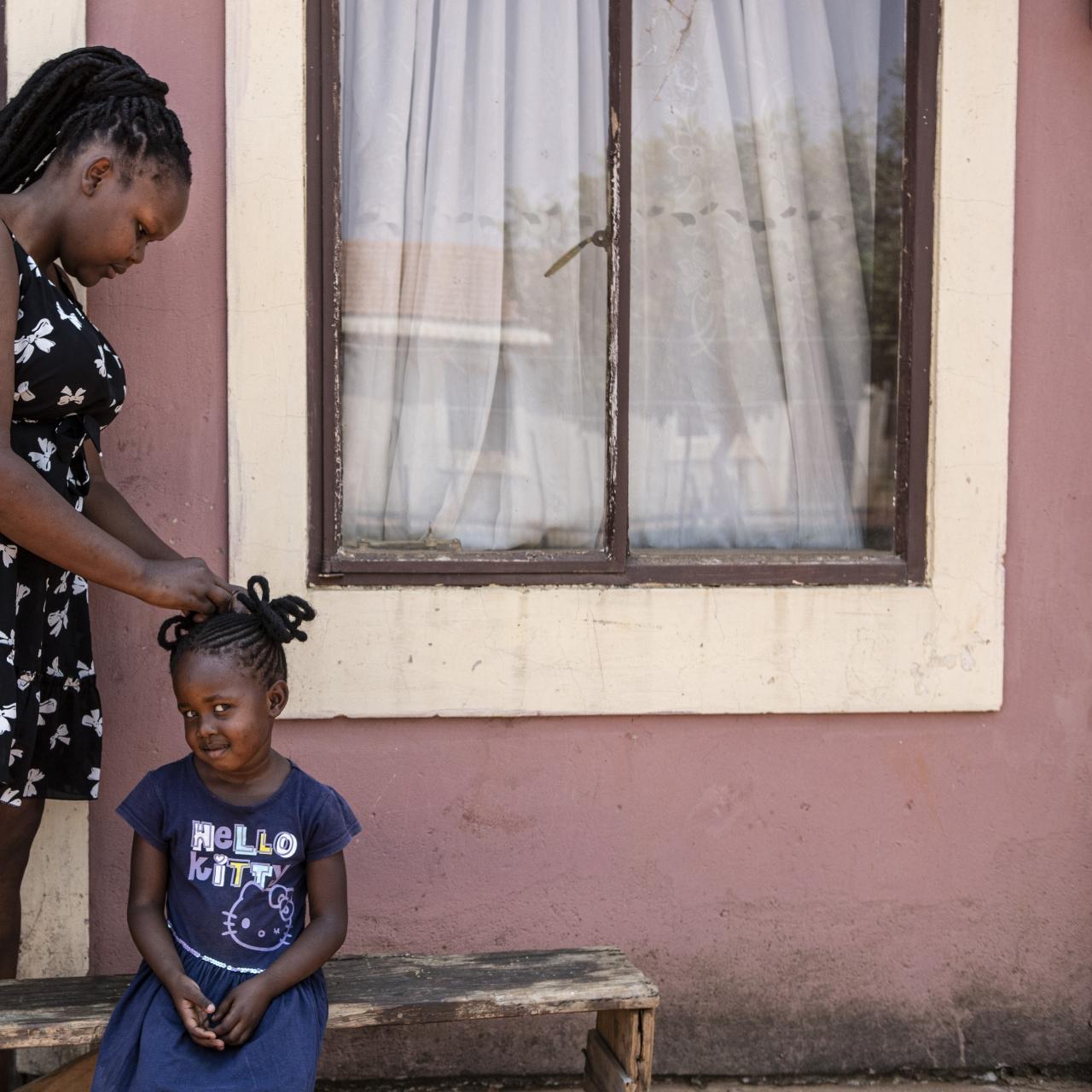 Xoli, a young woman in a black and white dress braiding the hair of a young girl (her sister) on the front step of a house