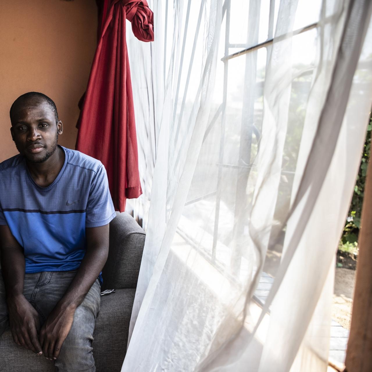Thabang, a young man in a blue t-shirt sits on a grey sofa next to a window in small orange room