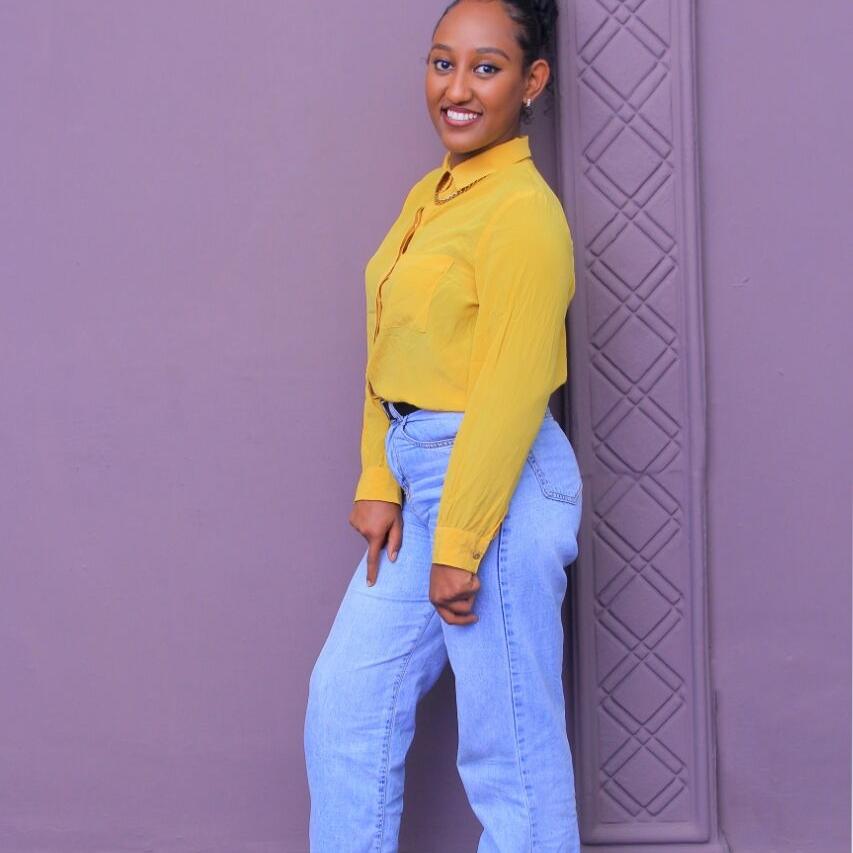 Beza, a young woman in a bright yellow jacket and jeans stands in front of a lilac wall