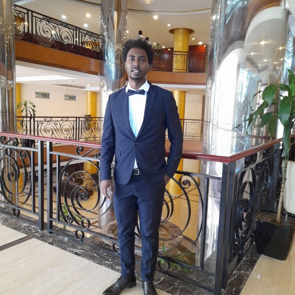 Habte, a young man in a smart black suit stands in front of a the railings around a hotel atrium