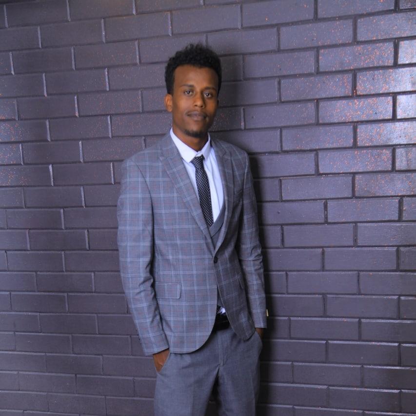 Henock, a young man in a smart grey suit stands in front of a shiny black brick wall