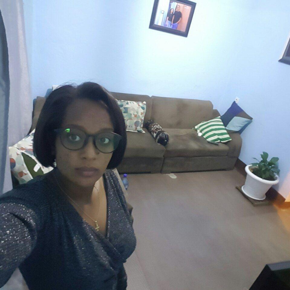 A selfie of a woman standing in the corner of a living room with white walls, brown sofa and pictures hanging on the wall
