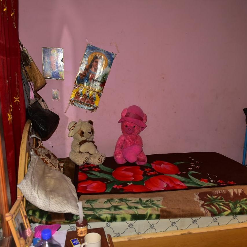 A small pink and turquoise bedroom with with religious posters on the wall above a small bed covered in decorative throws and teddy bears