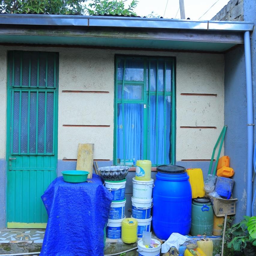 A small one-storey house with white and blue walls, a green door and windows with various containers stacked up against the wall beneath a window