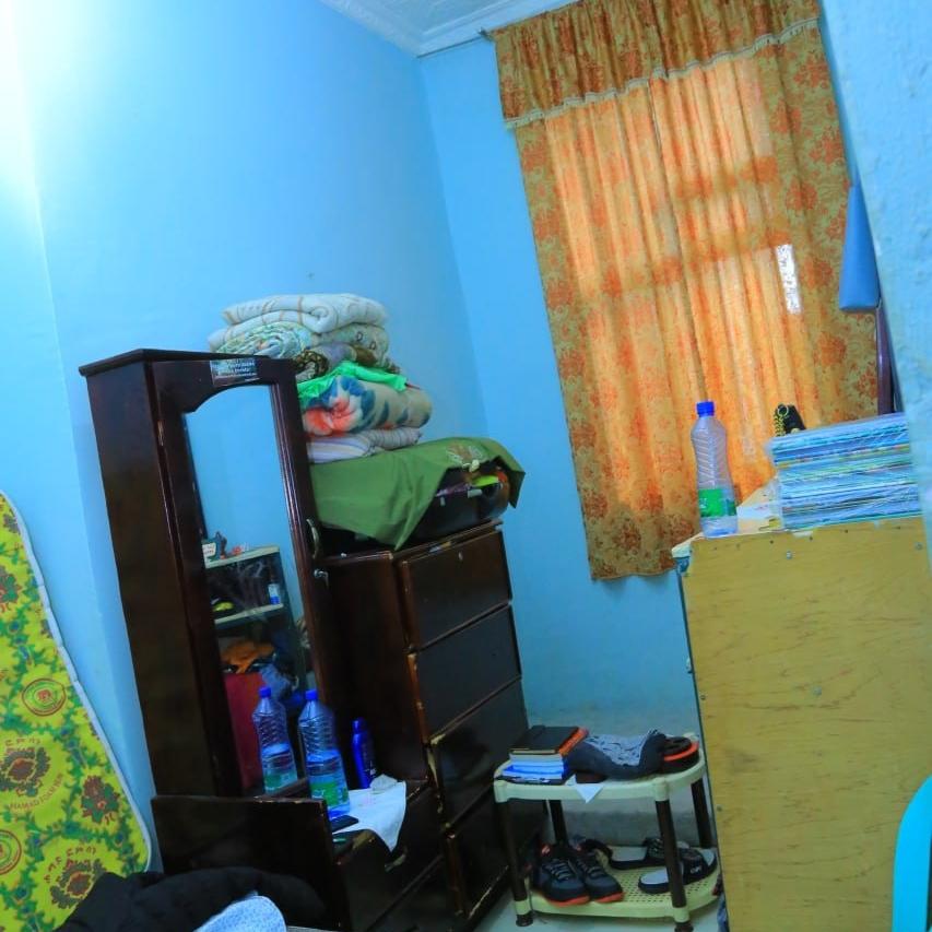 A small cluttered bedroom with light blue walls, patterned orange curtains and dark wooden bedroom furniture