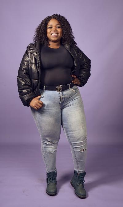 Tsholo, a young woman in a black PVC bomber jacket and jeans stands in front of a lilac wall