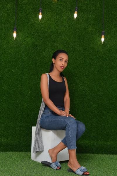 Zufan, a young women in a black vest, jeans and sandals sits on a white cube in front of a wall of grass