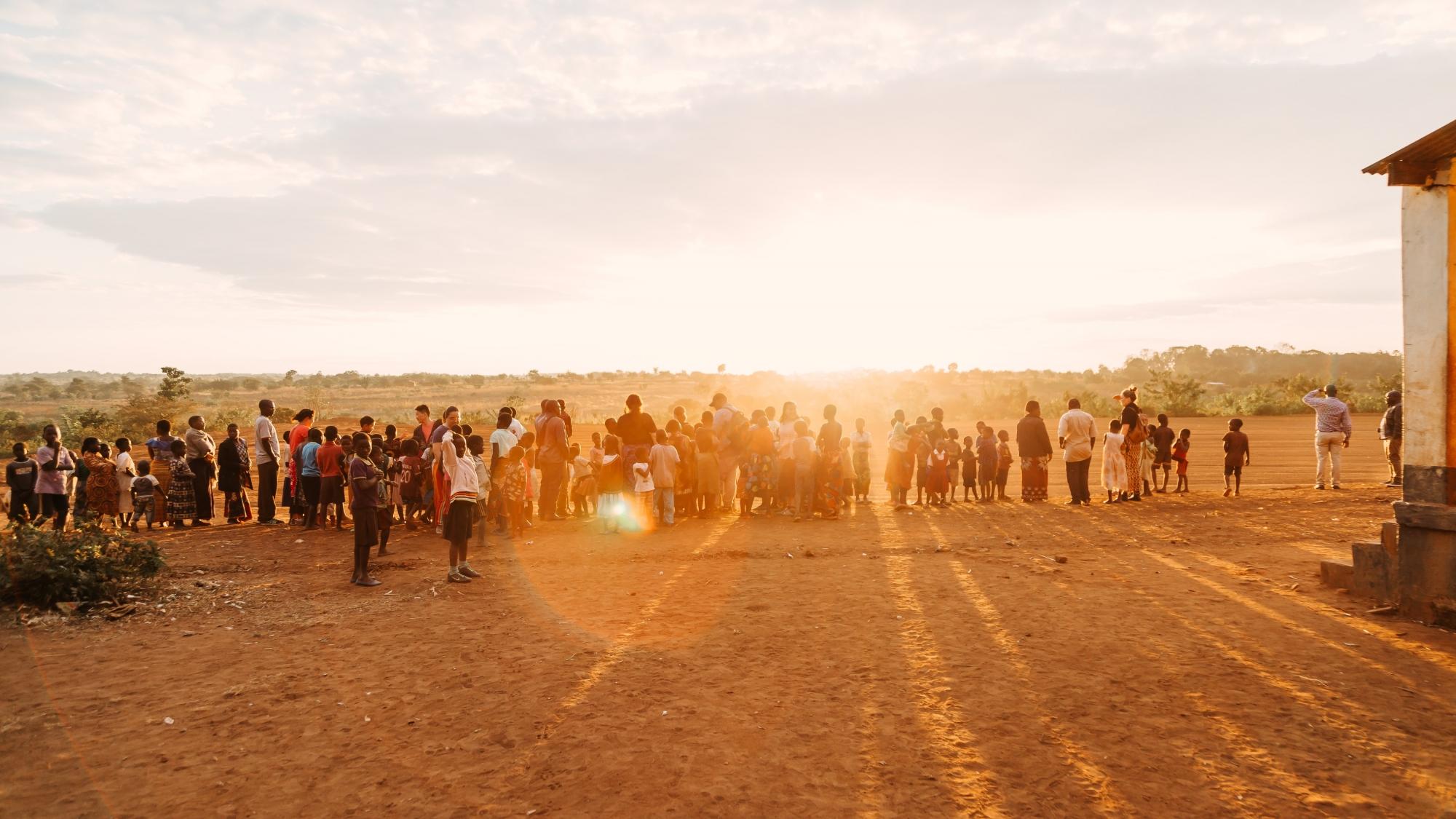 Adults and children stand in front of sun that is setting on horizon and casting a golden light over the foreground