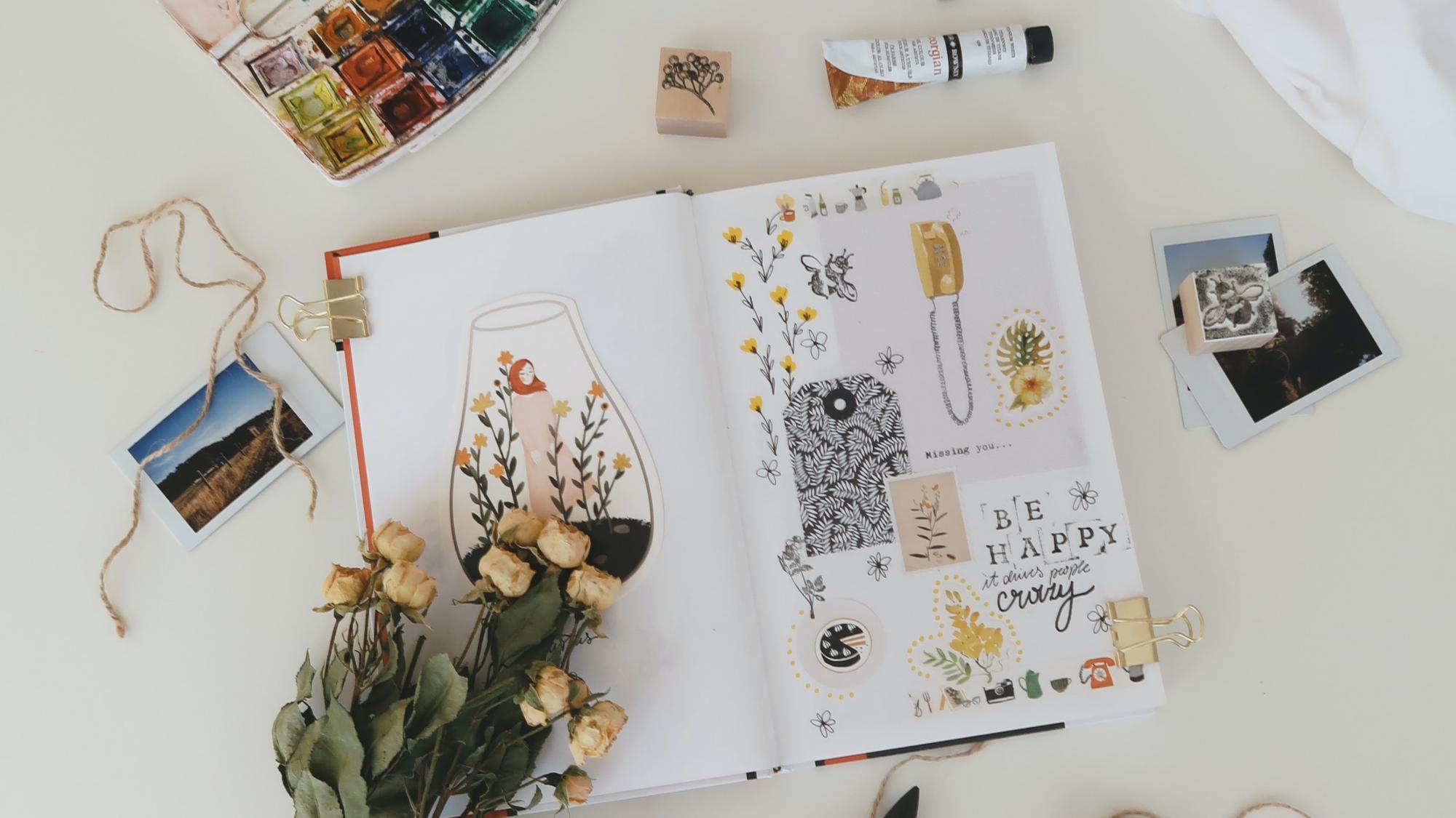 A journal, paints, pens, flowers and photos lay scattered aesthetically on a table