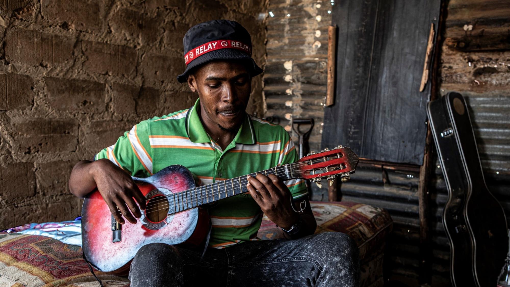 A young man sits on a mattress in a small shack, playing the guitar