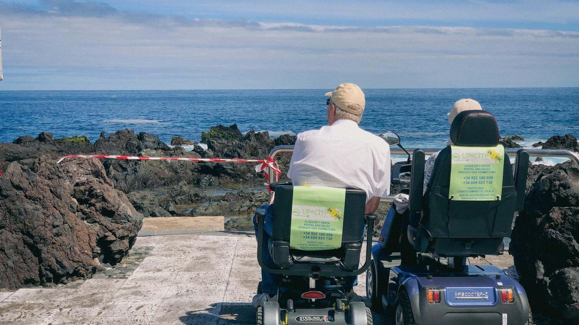 two elderly people side by side sat on mobility scooters at the Spanish coast