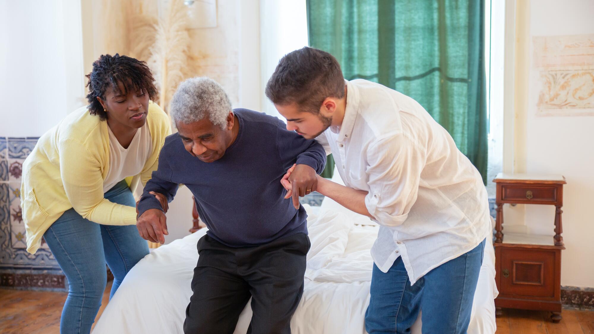 A man and woman helping an elderly man to stand up from a bed
