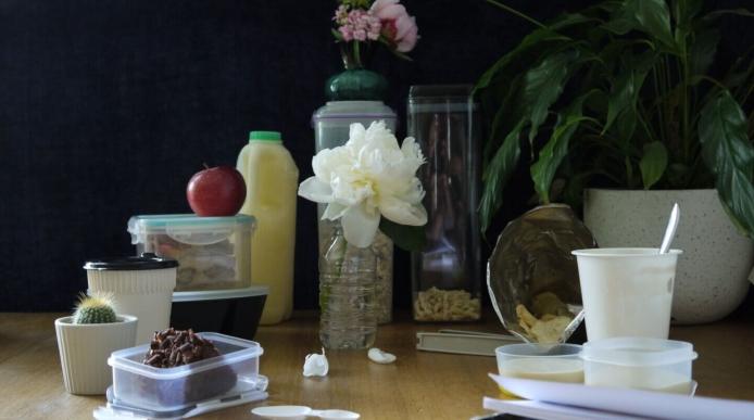 Household items made out of plastic placed on a table top, including a tupperware container, a plastic bottle and a yoghurt pot.