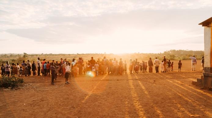 Adults and children stand in front of sun that is setting on horizon and casting a golden light over the foreground