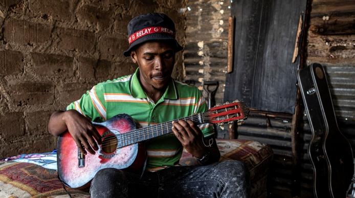 A young man sits on a mattress in a small shack, playing the guitar