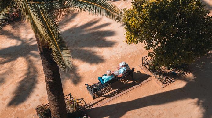 Two elderly people sat side by side on a bench under a palm tree
