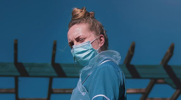 A healthcare worker in blue uniform, wearing a PPE mask and looking down at the camera