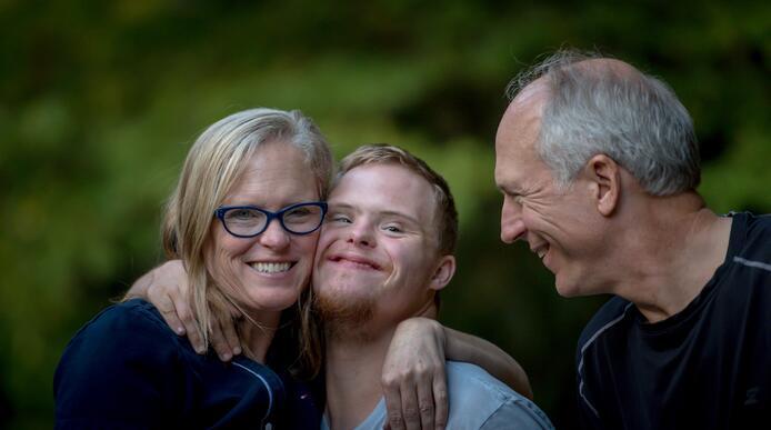 A young adult with Down's Syndrome smiling with his arm around an older woman and an older man stood beside him 
