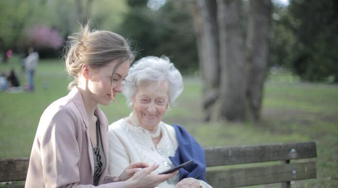 An elderly woman with younger woman sat on a bench in a park looking at a phone together 