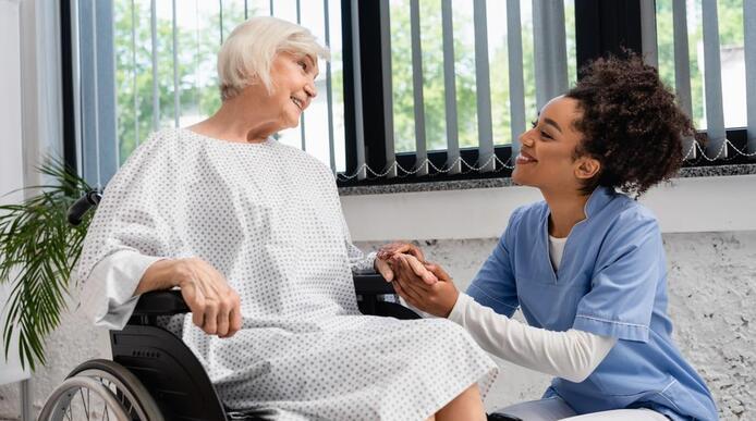An elderly lady in a wheelchair holding hands with a caregiver who crouches next to her