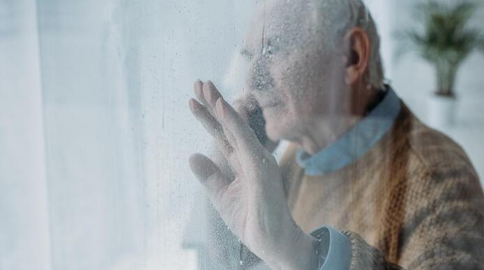 An elderly man looking out the window with one hand against the glass