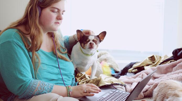 A woman using a laptop sits on a bed next to a small dog
